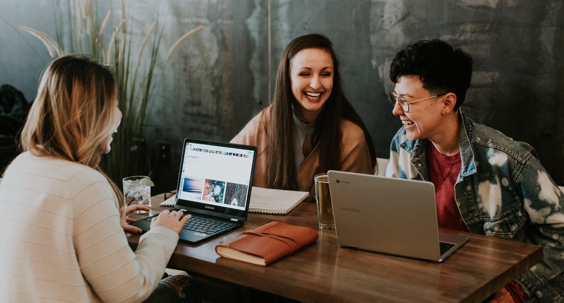 Three people wearing casual business attire laugh together as they work on laptops in coffee bar.