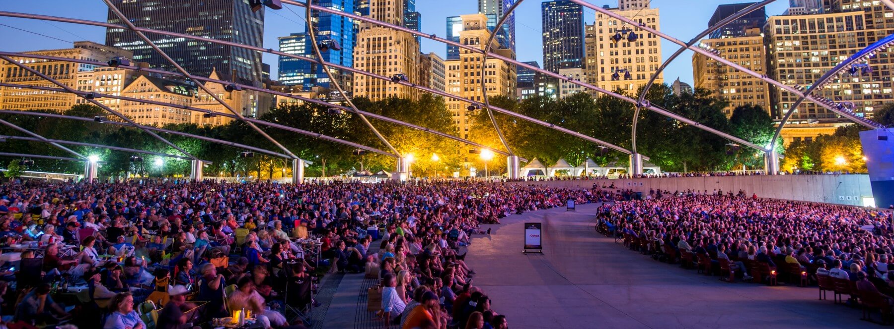 An audience watches a performance take place on the Jay Pritzker Pavilion, in front of the Chicago skyline.